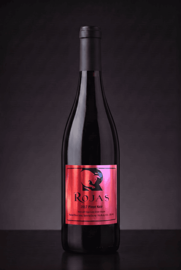A dark glass wine bottle with a shiny red label reading Rojas 2017 Pinot Noir stands upright against a dark, textured background.