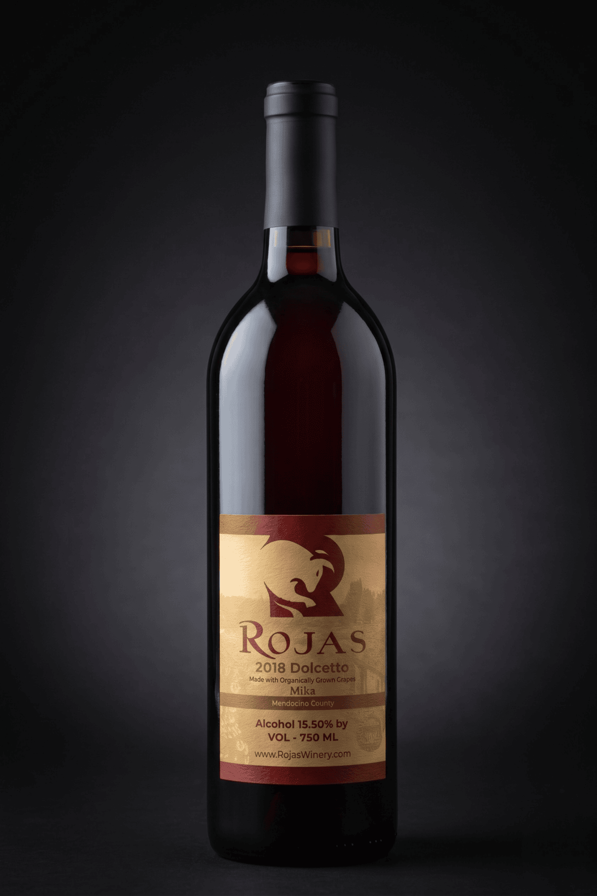 A bottle of red wine labeled Rojas 2018 Dolcetto with gold and brown tones, standing upright against a dark background.