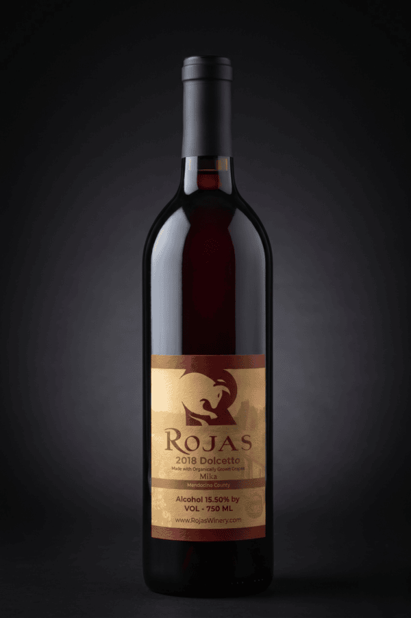 A bottle of red wine labeled Rojas 2018 Dolcetto with gold and brown tones, standing upright against a dark background.