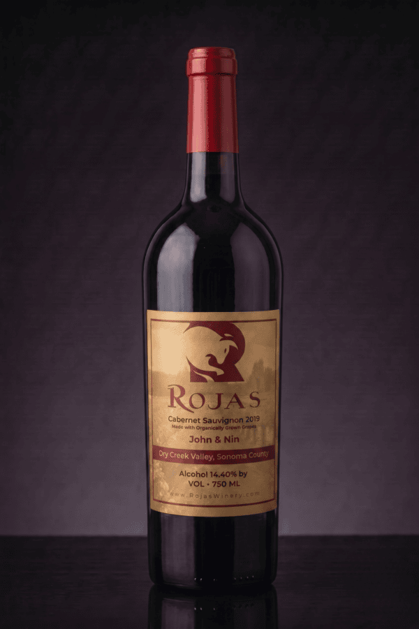 A bottle of Rojas Cabernet Sauvignon 2019 wine with a red cap and gold label, photographed against a dark, textured background.