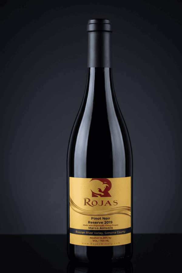A bottle of Rojas Pinot Noir Reserve 2019 wine stands upright against a dark background, featuring a gold label with black and white text and a stylized bird logo.