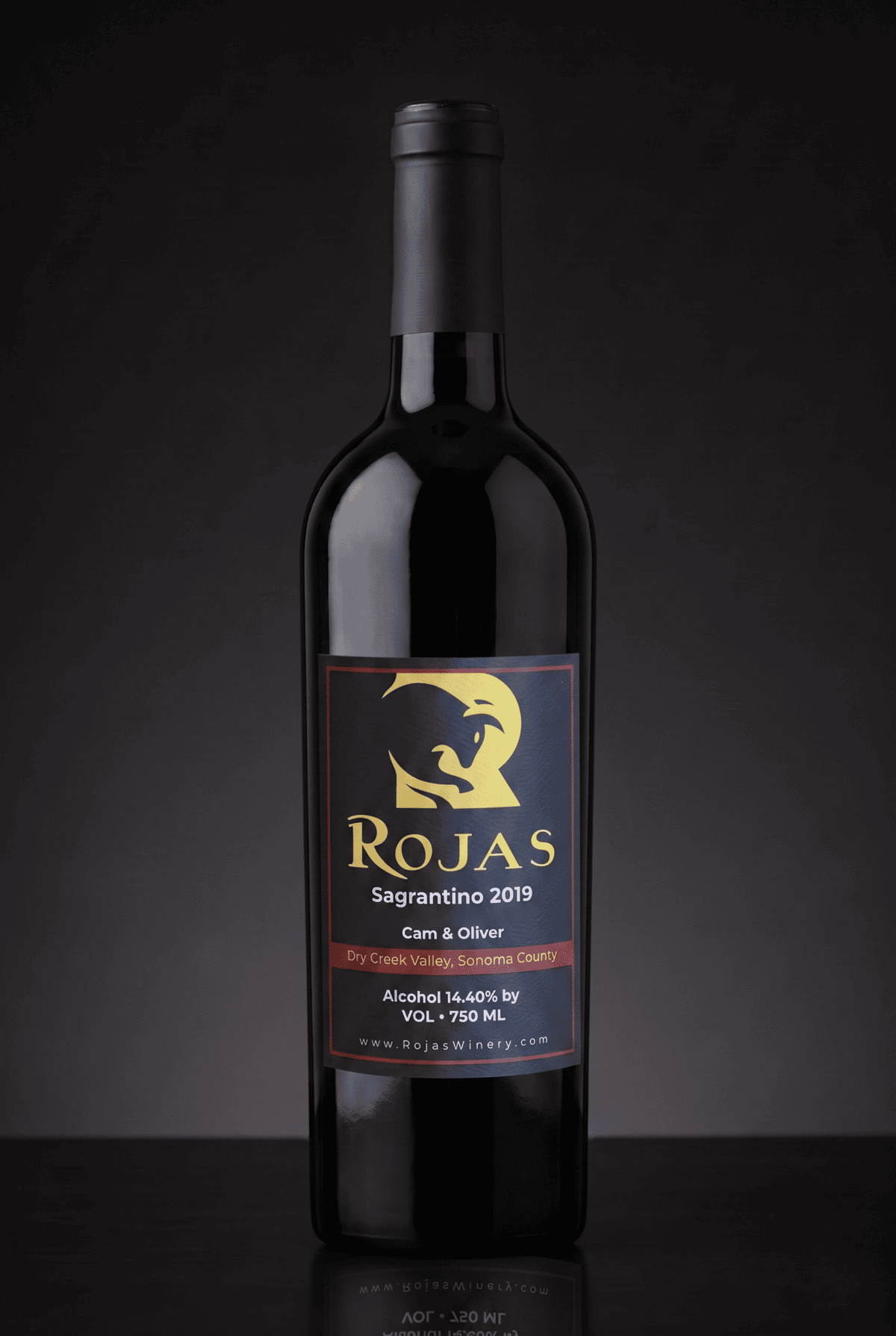 A bottle of Rojas Sagrantino 2019 red wine stands on a dark surface, with a gold and red label featuring a stylized animal and winery details. The background is black and softly lit.