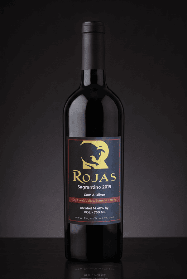 A bottle of Rojas Sagrantino 2019 red wine stands on a dark surface, with a gold and red label featuring a stylized animal and winery details. The background is black and softly lit.