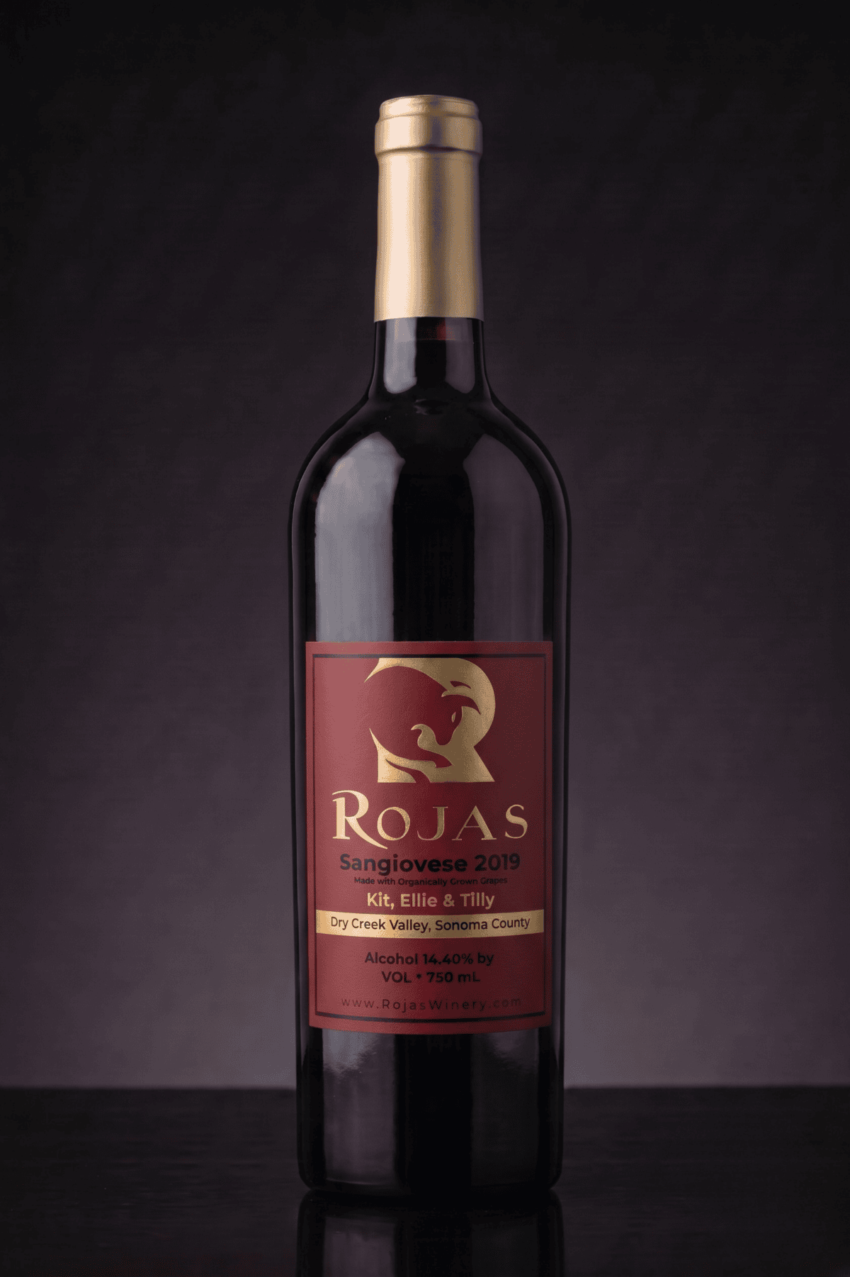A bottle of Rojas Sangiovese 2019 wine with a gold cap and a red and gold label, standing upright against a dark gradient background. The label details the wines origin and alcohol content.