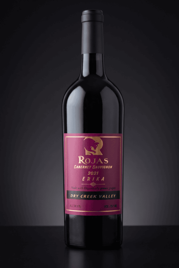 A bottle of Rojas Cabernet Sauvignon 2021 wine with a purple and gold label, set against a dark gradient background. The label highlights Dry Creek Valley and Made with organically grown grapes..