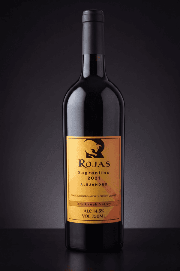 A bottle of Rojas Sagrantino 2021 red wine with a gold label, featuring Alejandro, Dry Creek Valley, 14.5% alcohol, and 750ml size, set against a dark background.