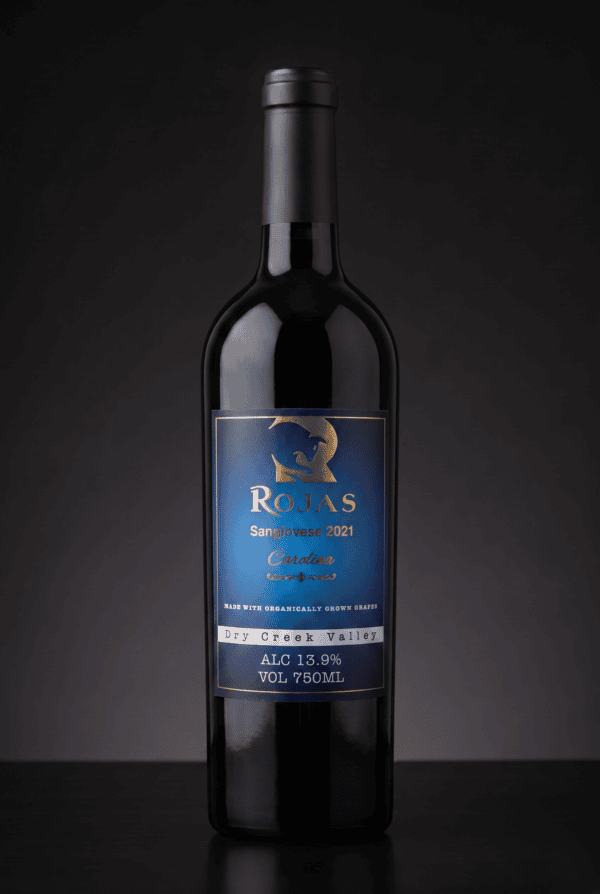 A bottle of Rojas Sangiovese 2021 wine with a blue and gold label on a dark background. The label shows alcohol content 13.9% and volume 750ml.