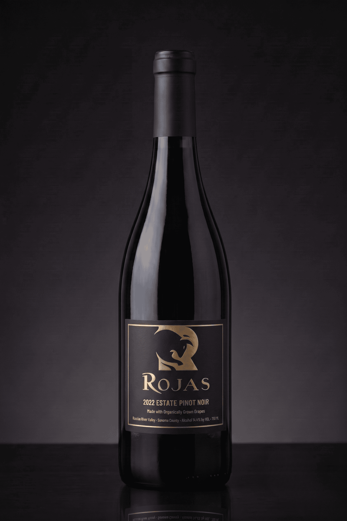 A dark glass wine bottle labeled Rojas 2022 Estate Pinot Noir stands upright against a dark, gradient background. The label features gold text and a stylized bird logo.