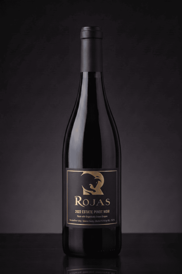 A dark glass wine bottle labeled Rojas 2022 Estate Pinot Noir stands upright against a dark, gradient background. The label features gold text and a stylized bird logo.
