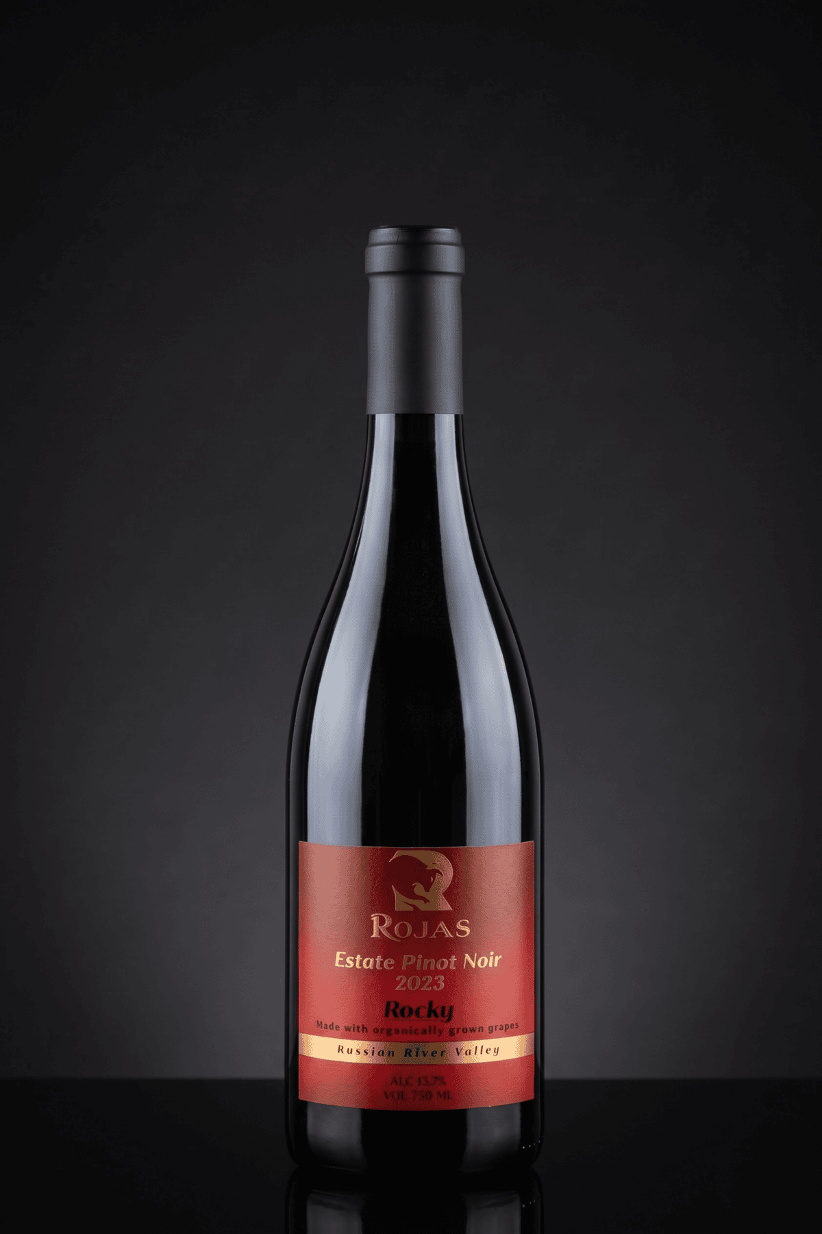 A bottle of Rojas Estate Pinot Noir 2023 wine with a red and gold label stands upright on a dark reflective surface against a black background.