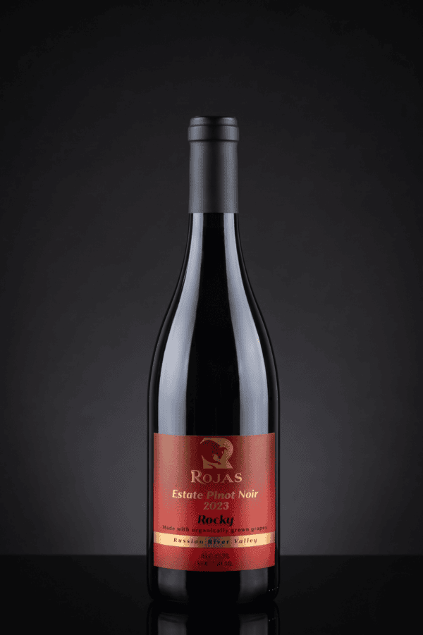 A bottle of Rojas Estate Pinot Noir 2023 wine with a red and gold label stands upright on a dark reflective surface against a black background.