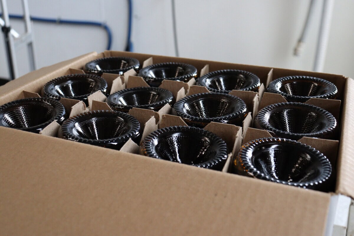 A cardboard box holds 15 black glass bottles placed upside down, separated by cardboard dividers. Blue pipes and a white wall are visible in the blurry background.