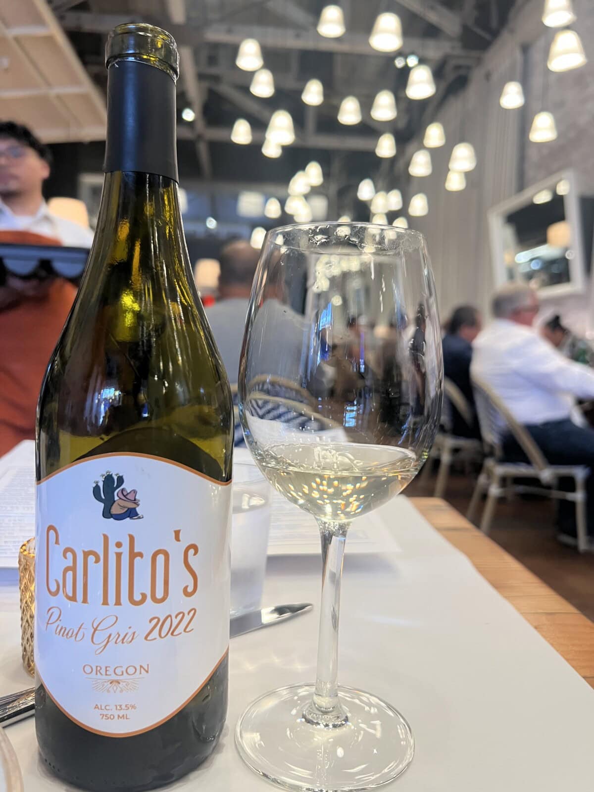 A bottle of Carlitos Pinot Gris 2022 wine and a half-filled wine glass sit on a restaurant table, with blurred diners and hanging lights visible in the background.