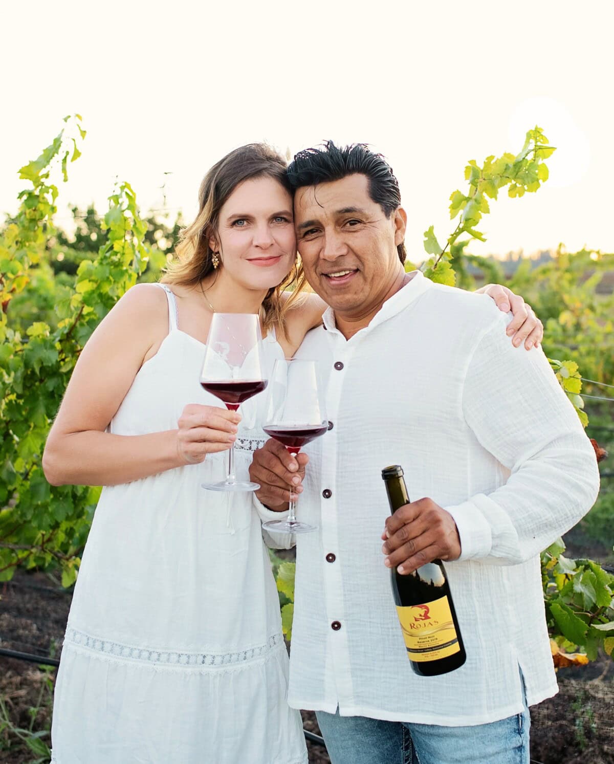 A woman and a man stand close together in a vineyard, holding glasses of red wine. The man also holds a wine bottle. Both are dressed in white and smiling at the camera, with green grapevines in the background.