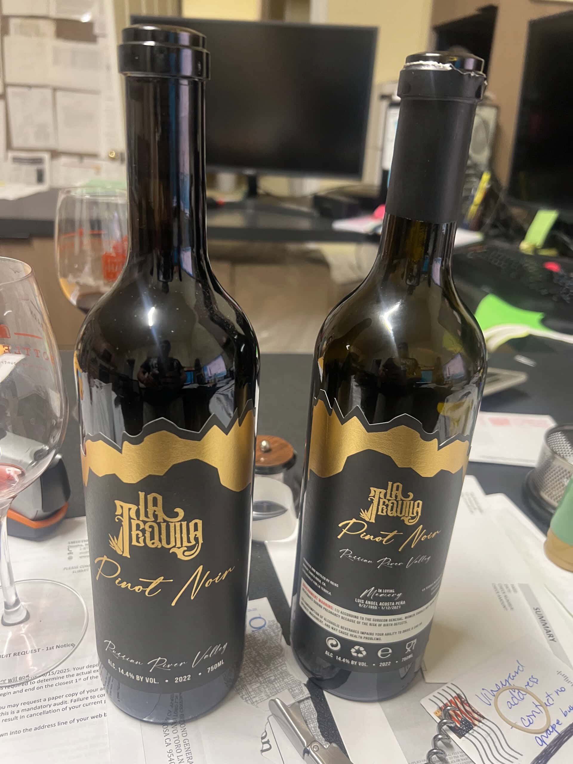Two bottles of La Tava Pinot Noir wine are on a cluttered desk with papers, a wine glass, and computer monitors in the background. Both bottles have black and gold labels.