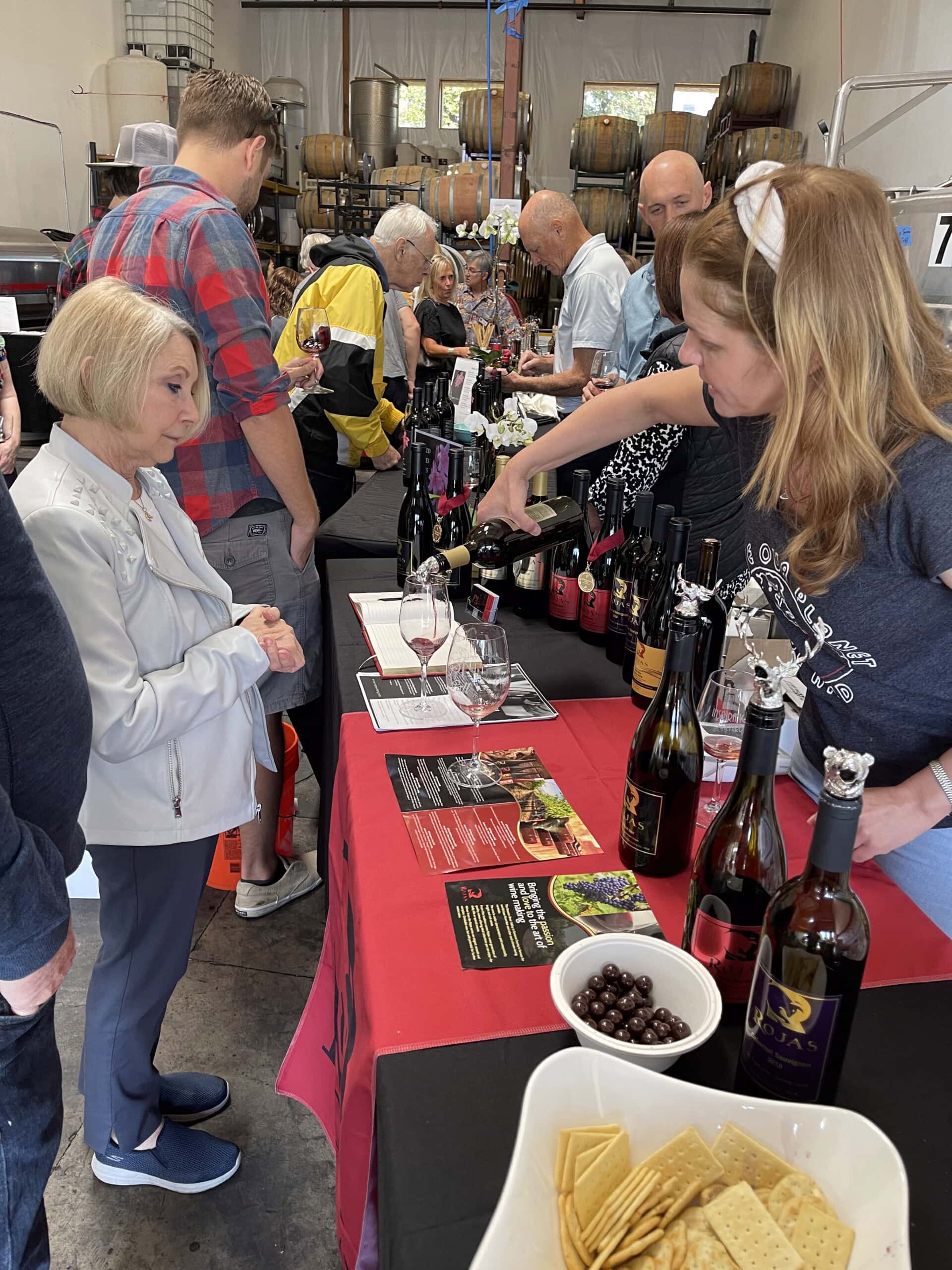 A woman pours wine for a guest at a busy wine tasting event. Wine bottles, glasses, snacks, and pamphlets are on the table. People wait in line in a casual indoor setting with barrels in the background.