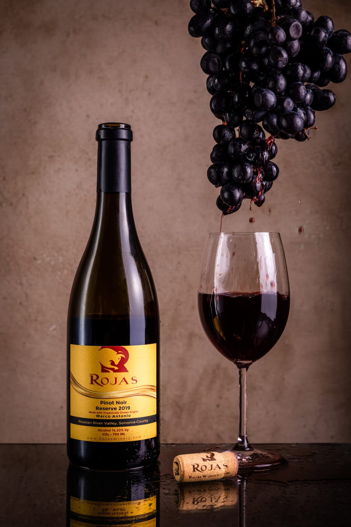 A bottle of Rojas Pinot Noir Reserve 2019, a cork, a glass of red wine, and a bunch of grapes dripping juice into the glass, set against a brown background.