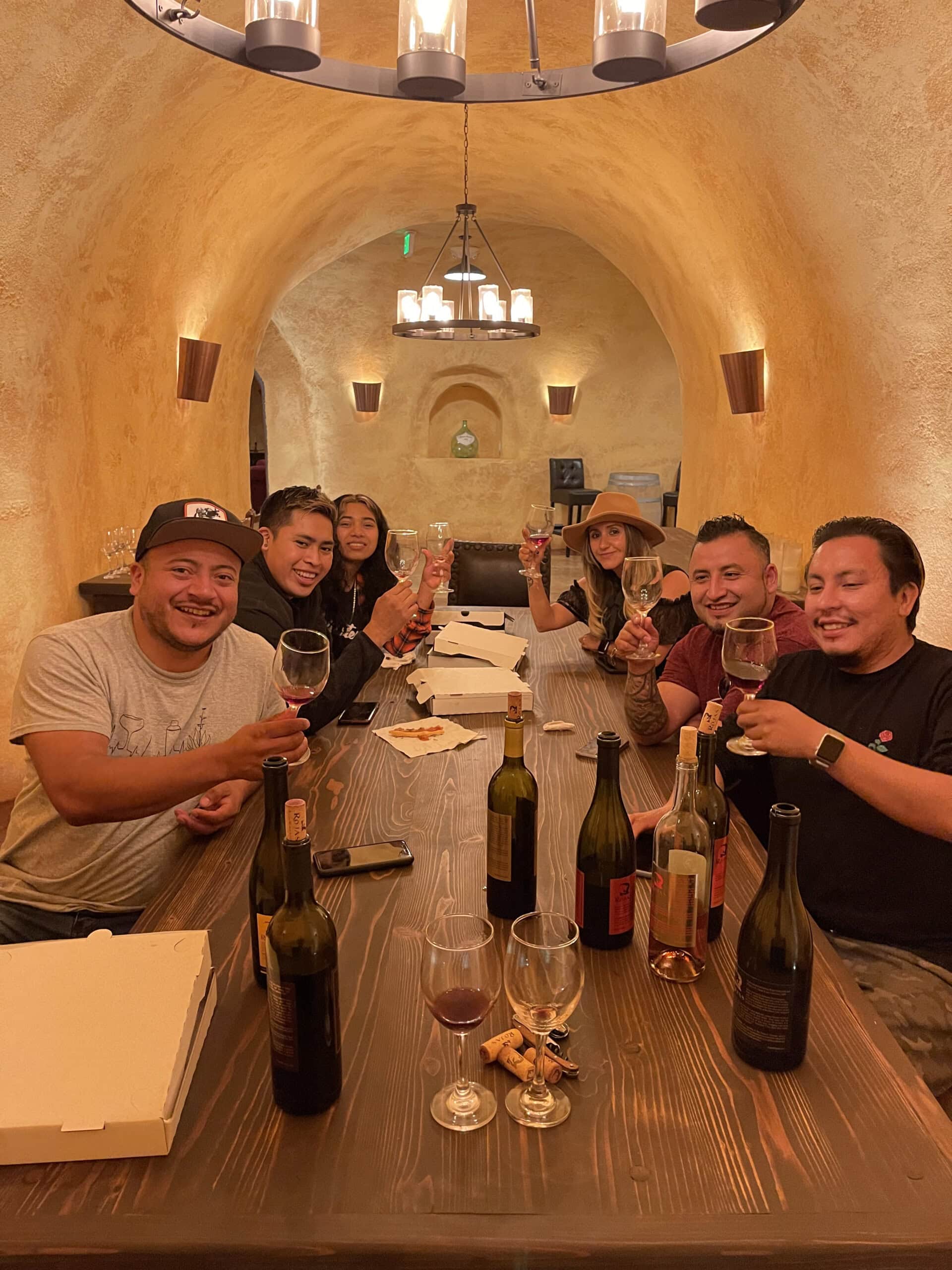 Six people sit at a long wooden table in a cozy, cave-like room, smiling and raising wine glasses. Wine bottles, glasses, and pizza boxes are on the table under warm lighting.