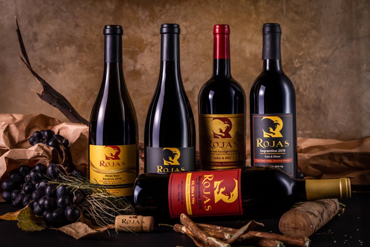 Five Rojas wine bottles, four standing and one lying down, are displayed with grapes, corks, and dried leaves on a rustic surface. The labels are black, gold, and red, each showing a griffin-like logo.