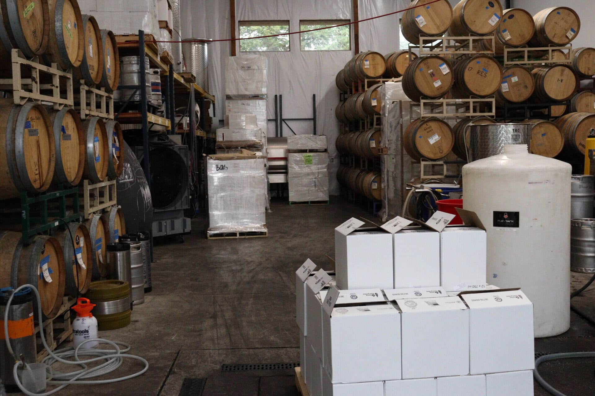 A storage room filled with wooden barrels stacked on racks, white cardboard boxes in the foreground, and a large white tank on the right. Shelves and pallets with packaging materials are visible in the background.