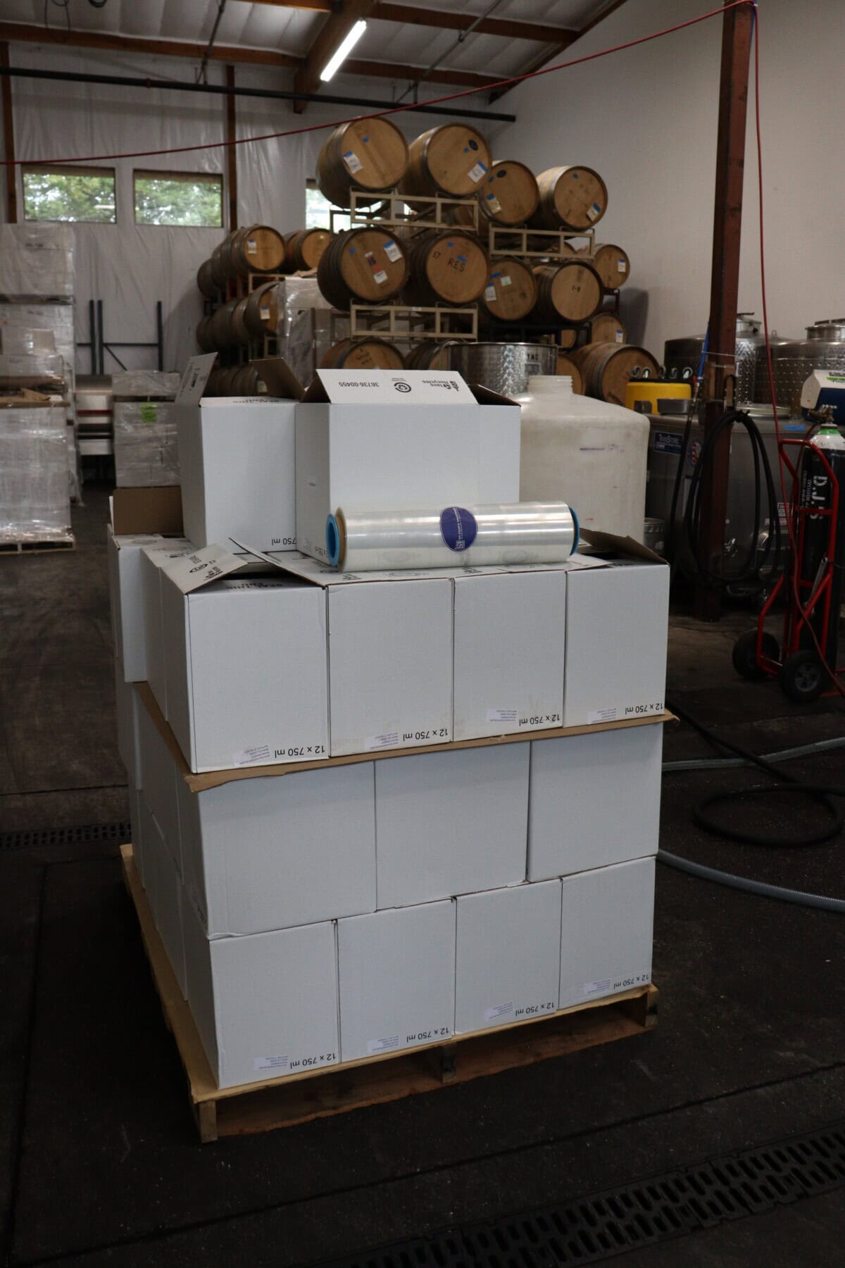 A pallet stacked with white boxes and a roll of plastic wrap sits in a warehouse. In the background, there are barrels, tanks, and industrial equipment.