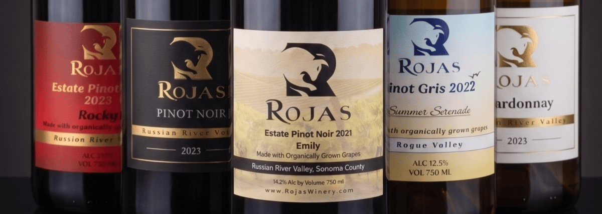 Five bottles of Rojas wine are lined up, each labeled with different varietals and years, including Pinot Noir, Pinot Gris, and Chardonnay, with details about regions and organic grapes.